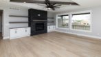 light blonde wood flooring, black stone fireplace, large windows, large beams with ceiling fan overhead