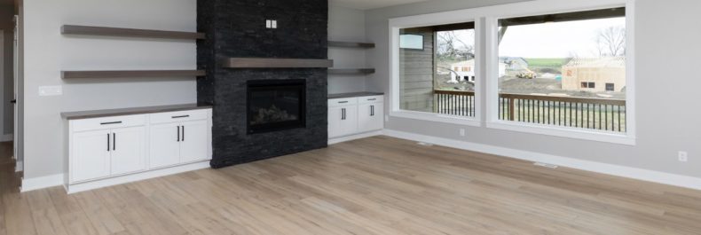 light blonde wood flooring, black stone fireplace, large windows, large beams with ceiling fan overhead