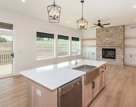 Black House Kitchen Island and Fireplace open concept kitchen and living area with light wood plank flooring, light wood cabinet island, white countertops, large wall of open windows and stacked stone fireplace