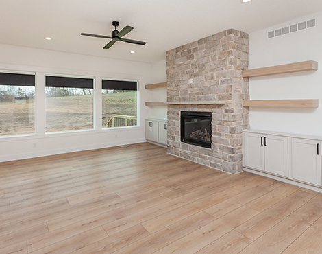 Black House Living Room living area with light wood plank flooring, stacked stone fireplace, taupe cabinets with light wood floating shelves above and large open windows