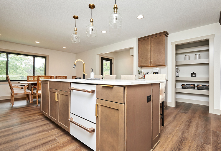 light wood cabinets, and white dishwasher with gold hardware, glass and gold hanging pendant lights looking into pantry